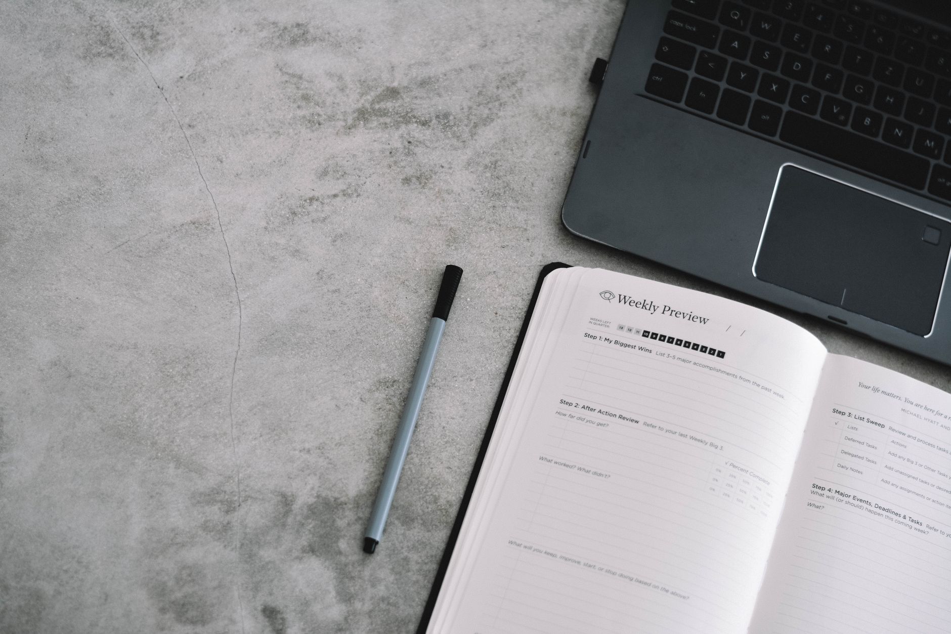A person sits at a desk reviewing a paper weekly planner next to a laptop showing an AI assistant.