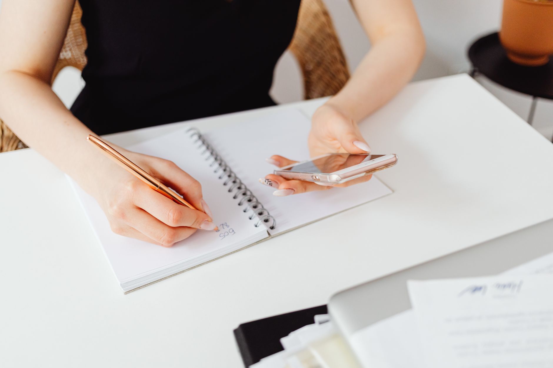 Hands jotting social media post ideas in a notebook while a phone shows a content calendar.