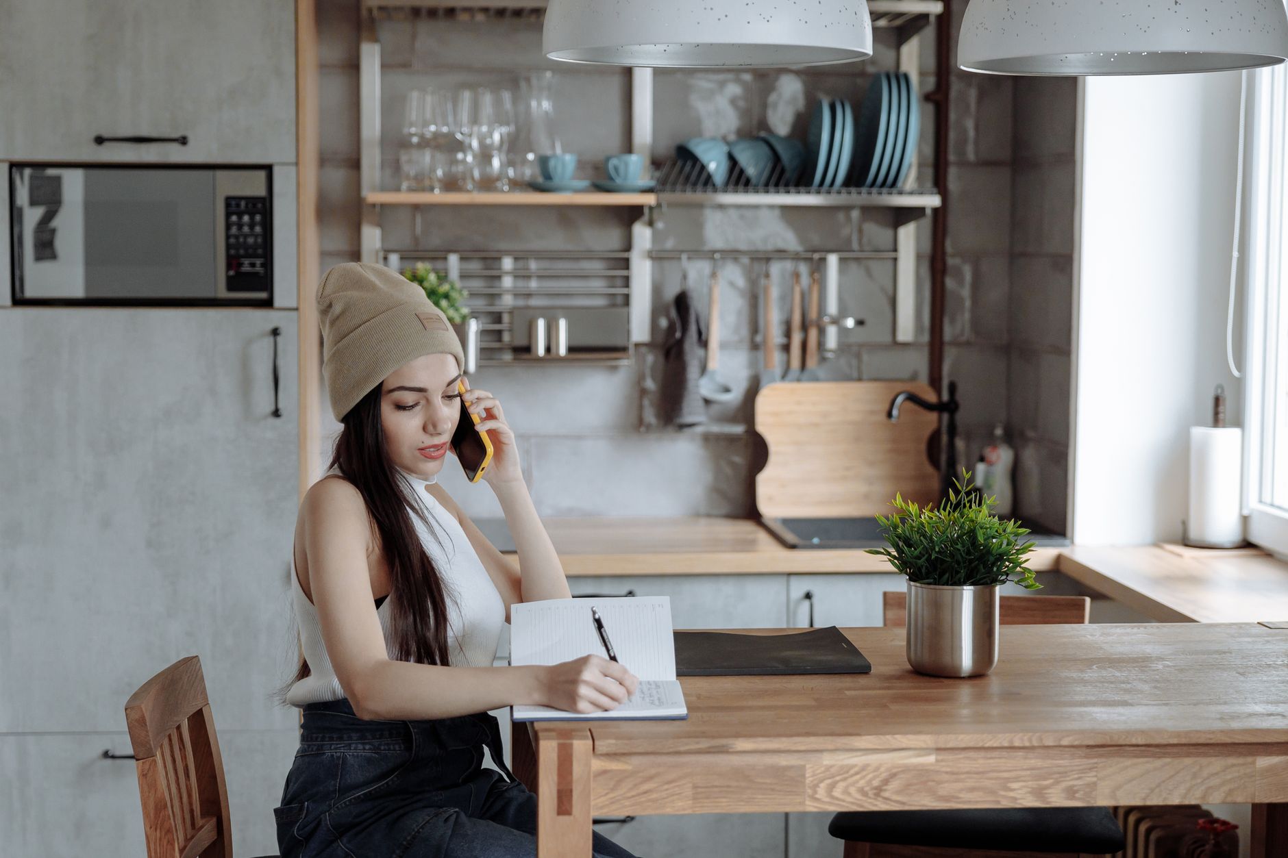 A woman writes in a notebook at a bright kitchen table while a smartphone and laptop are nearby.